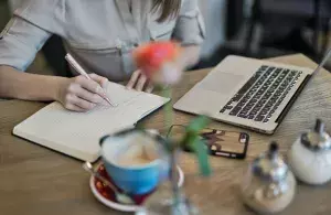 Das Bild zeigt eine Person am Laptop mit Notizbuch und Kaffeetasse.