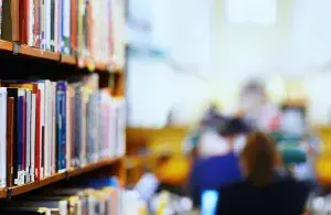 Eine Reihe Bücher in einer Bibliothek, im Hintergrund sind Lesende an Tischen zu sehen.