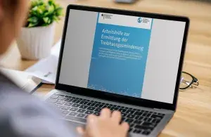 Person arbeitet an einem Laptop, auf dem ein Dokument mit dem Titel "Arbeitshilfe zur Ermittlung der Treibhausgasminderung" angezeigt wird. Das Dokument stammt vom Bundesministerium für Wirtschaft und Klimaschutz und der Nationalen Klimaschutzinitiative.