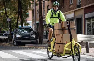 Fahrer mit Warnjacke transportiert Pakete auf einem Lastenrad durch eine Stadtstraße.