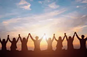 Menschen stehen im Sonnenuntergang nebeneinander und halten einander die erhobenen Hände.