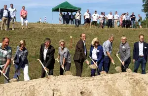 Eine Gruppe von Menschen setzt Spaten in einen Erdhügel, während weitere Personen den Spatenstich verfolgen.