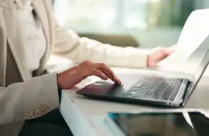 Eine Person bedient einen Laptop und hält in der anderen Hand ein Blatt Papier.