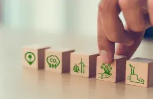 Auf dem Foto greift eine Hand nach einem Holzwürfel auf dem grüne Münzen aufgedruckt sind. Der Würfel liegt in einer Reihe von Holzwürfeln die alle Symbole rund um das Thema Klimaschutz und Bilanzierung tragen.