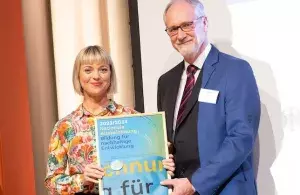 Nadja Kulikowa (links, WWF) und Dr. Klaus Grosfeld (rechts, REKLIM) stehen auf der Bühne und halten die Urkunde für die „Nationale Auszeichnung – Bildung für nachhaltige Entwicklung“ in den Händen. Beide blicken freundlich in die Kamera.