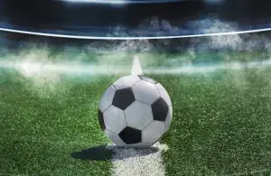 Ein Fußball liegt auf dem Anstoßpunkt eines Spielfelds in einem beleuchteten Stadion unter Flutlicht, im Hintergrund Nebelschwaden.