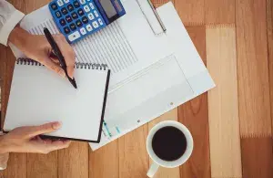 Zu sehen sind auf dem Foto eine Schreibtischansicht von oben. Auf dem Tisch steht eine Kaffeetasse und eine Person hält einen Block und Stift. Daneben liegt ein Taschenrechner und weitere Unterlagen. 