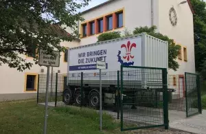 Ein LKW-Anhänger steht vor einem Haus, drumherum ein Zaun