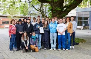 Gruppe Jugendlicher und Erwachsener steht unter einem Baum im Schulhof, eine Person hält eine Urkunde.