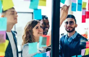 Das Foto zeigt ein diverses Team im Gespräch. Gemeinsam werden Post-Ins an eine Wand geklebt und darüber gesprochen. 