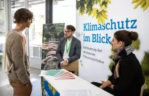 Zwei Mitarbeitende der Agentur für kommunalen Klimaschutz unterhalten sich an ihrem Informationsstand mit einer interessierten Person.