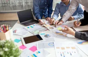 Eine Gruppe von Personen beugt sich über einen Tisch, auf dem ein Laptop, ein Tablet und mehrere Dokumente mit Bilanzen und Graphiken verteilt sind. Gemeinsam planen und diskutieren sie. Von den Personen sehen wir lediglich die Hände.  