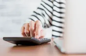 Eine Hand tippt auf einem Taschenrechner, im Vordergrund verschwommen ein Laptop