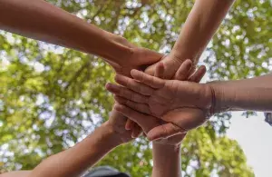 Mehrere Menschen halten ihre Hände in einem Kreis übereinander als Symbol des Zusammenhalts. Im Hintergrund sind Baumkronen zu erkennen. 