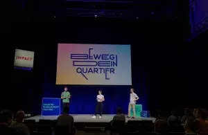 Drei Personen stehen auf einer Bühne. Im Hintergrund ein Bildschirm auf dem „Beweg dein Quartier“ steht.