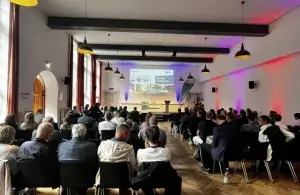 In einem Saal sitzen zahlreiche Menschen vor einer Leinwand mit einer Präsentation.