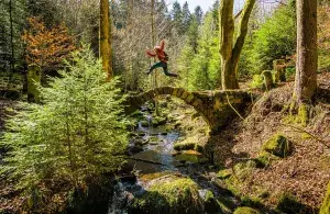 Eine Person springt auf einer Steinbrücke in die Höhe, die Brücke führt über einen Bach in idyllischer Umgebung.
