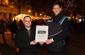 Thomas Fischer, Leiter Kreislaufwirtschaft Deutsche Umwelthilfe und Martin Hikel, Bezirksbürgermeister von Berlin-Neukölln, stehen nebeneinander und halten eine gerahmte Urkunde in der Hand. Martin Hikel hat ein Baby in einem Tragerucksack vor der Brust. Im Hintergrund ist der Alt-Rixdorfer Weihnachtsmarkt zu sehen. 